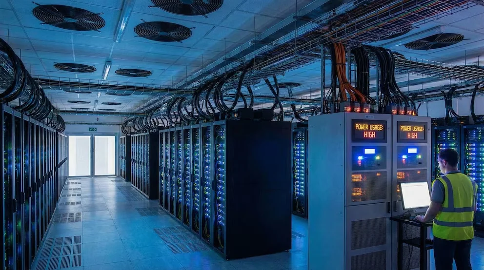Un technicien en gilet de haute visibilité supervise des rangées de serveurs avec des lumières bleues dans un centre de données. Des panneaux indiquent 'POWER USAGE: HIGH'.