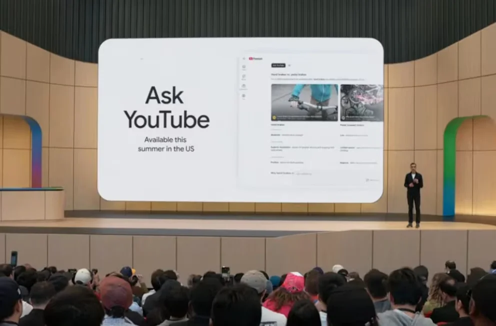Sundar Pichai debout sur une scène devant un grand écran affichant 'Demandez à YouTube' et des résultats de recherche, avec un public visible au premier plan