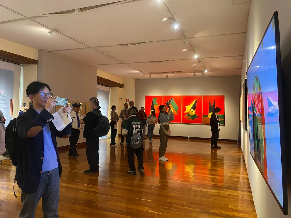 Plusieurs personnes dans une salle d'exposition d'art, observant des tableaux de fond rouge et des figures abstraites, et un écran numérique. Il y a un sol en bois et des lumières au plafond