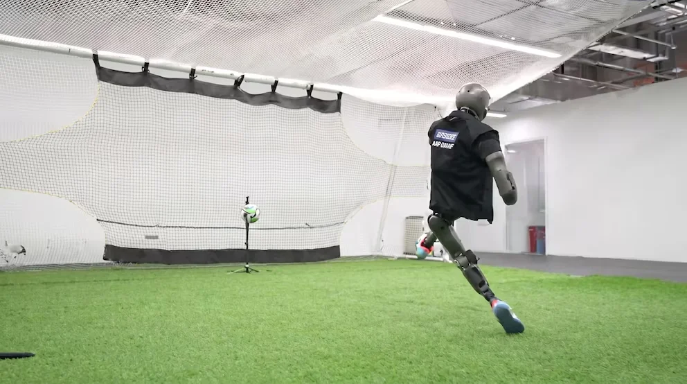 Un robot avec un gilet noir et des jambes articulées court sur un terrain de gazon synthétique vers un ballon de football, avec un but en arrière-plan.