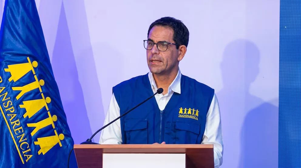Homme d'âge moyen portant des lunettes et un gilet bleu avec le logo de Transparence, parlant à un podium avec un microphone et un drapeau bleu à sa gauche.