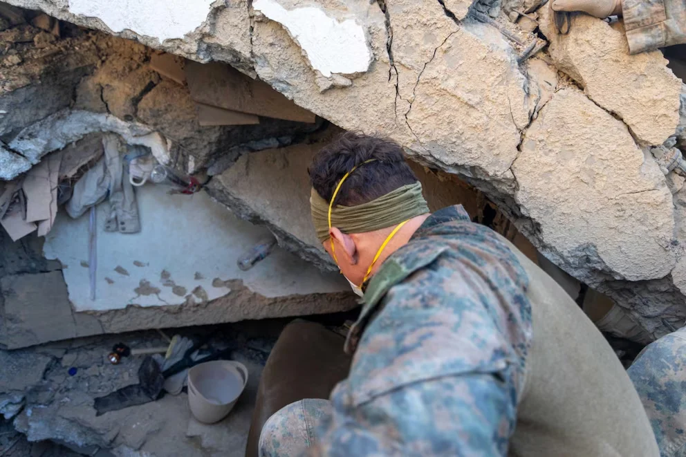 Personne en uniforme camouflé, masquée et bandana, s'accroupissant en observant parmi les décombres de béton cassé. On voit des morceaux de vêtements, des bouteilles et un seau.
