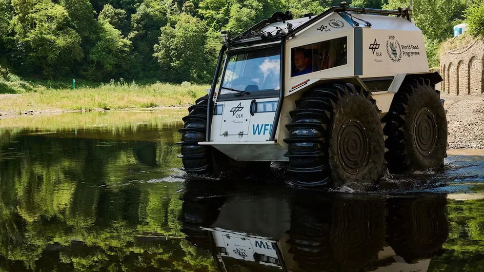 Véhicule amphibie Sherp blanc avec de grandes roues dans un corps d'eau. Logos DLR et Programme Mondial de l'Alimentation. Conducteur visible. Fond : arbres