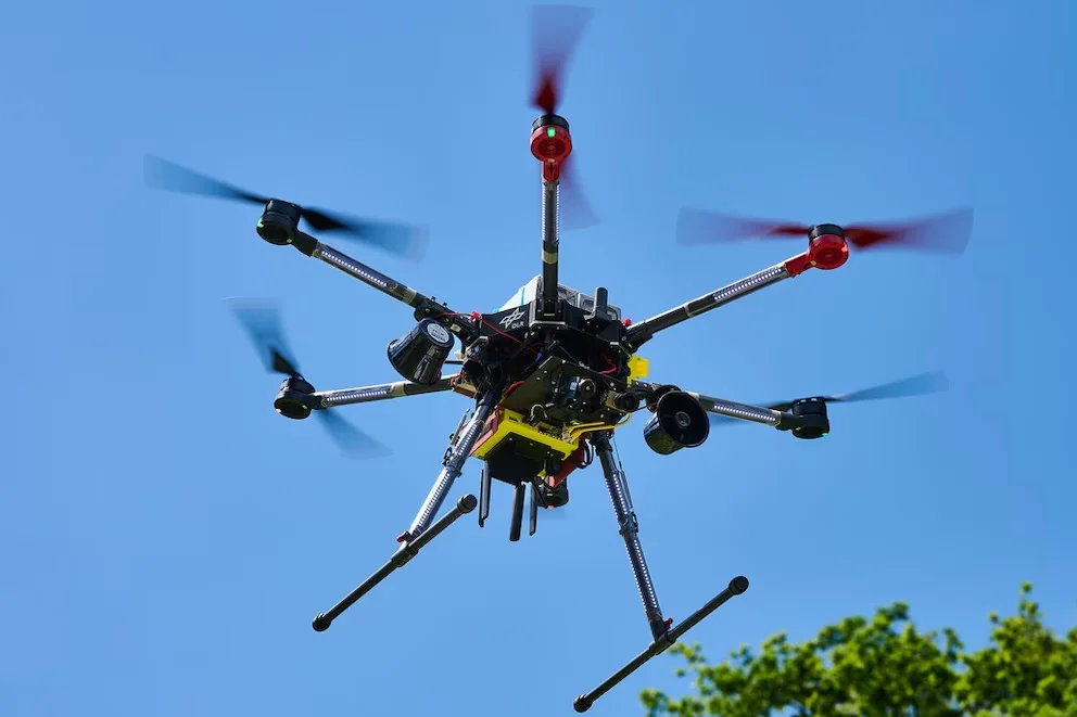 Drone hexacoptère noir avec six rotors en mouvement, quatre noirs et deux rouges, et train d'atterrissage métallique, suspendu dans un ciel bleu brillant