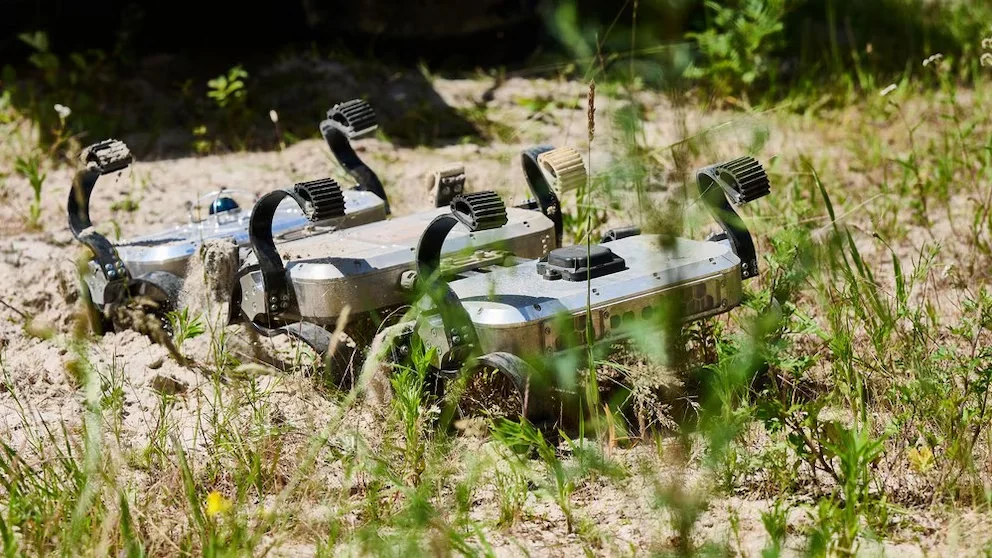 Gros plan d'un robot à quatre modules et multiples pattes ou roues métalliques sur un sol sablonneux avec de la végétation verte. Il y a une ombre partielle
