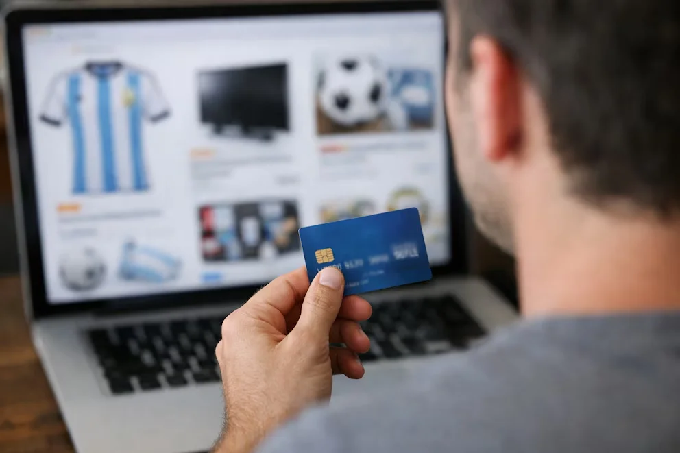 Un homme tient une carte de crédit d'une main devant l'écran flou d'un ordinateur portable qui affiche des articles en vente en ligne.
